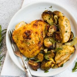 Golden and crispy rosemary chicken thighs with honey mustard apples and Brussels sprouts on a white plate with fresh rosemary on top.