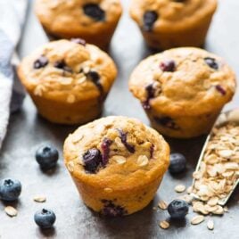 5 healthy blueberry muffins on a granite counter next to a scoop of rolled oats and fresh blueberries